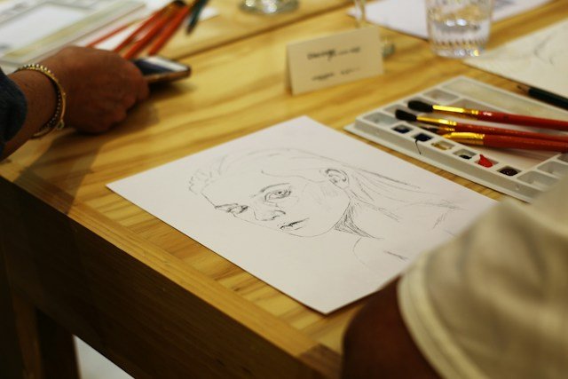 Pencil sketch of a human face portrait on a wooden table with paintbrushes and paints.